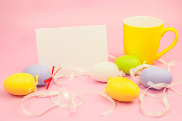 Easter composition with a white sheet of paper for writing text. Bright yellow cup on a pink background with colorful eggs, satin ribbons, white background with copy space