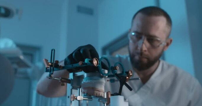 In the dark dental laboratory in the desk laying an axiosplit device inside with the ceramic model of upper jaw inside and male medical worker sitting near to the device and tight screw the details.