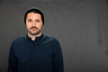 Handsome unshaven wimp wobbly guy in knitted cap posing on gray studio backdrop copy space. Male...