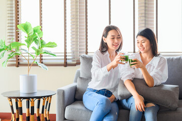 Two happy asian woman friends enjoy drinking green juice together in living room