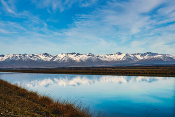 Snow covered Southern Alps mountain range near Twizel reflected in the still water in the canal flowing out of Lake Rautaniwha under some wispy cloud