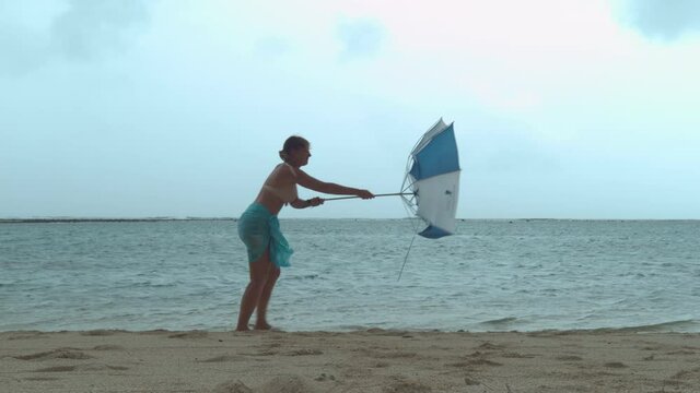 Young tourist struggles to hold her umbrella as she gets caught in a violent windstorm during her relaxing walk along the ocean. Funny shot of woman's umbrella getting destroyed by the powerful wind.