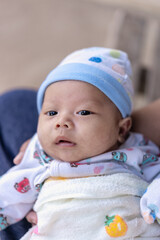 Selective focus on right eye of adorable newborn baby child boy resting in mother's arms at home. Headshot of cute Asian infant male looking at camera on mom's hands. New life and family concept.