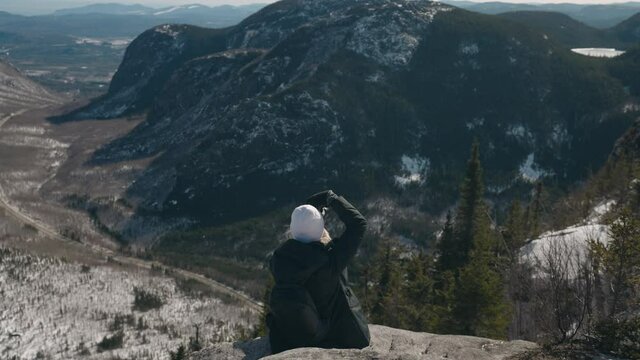 Girl Sitting On Edge Of Mountain Taking Photos At Mount of the Dome In Quebec, Canada. - rear, wide shot