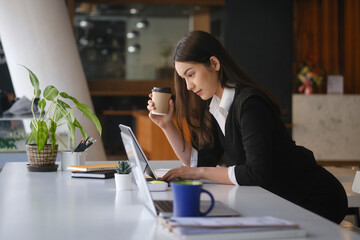Side view of businesswoman in suit holding cup of coffee and working with computer laptop at office.