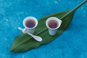 Two white cups with ceramic teaspoon with tea standing on a green leaf with blue background, view from top