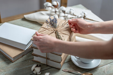 Women's hands is carefully decorating a stack of books with craft paper covers with rope and lavender flowers. Concept of packaging books for a gift