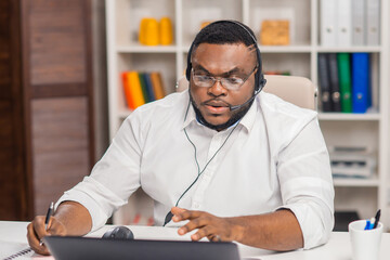 Workplace of freelancer. African-American man works at home office using computer, headset and other devices. Employee is having a conference call. Remote job.