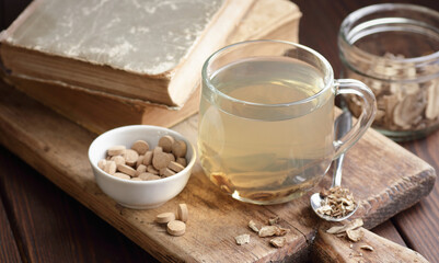 Burdock root tea in a glass cup with dry herb and old apothecary books nearby on wooden rustic background, closeup, naturopathy, natural medicine and homeopathy concept