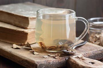 Burdock root tea in a glass cup with dry herb and old apothecary books nearby on wooden rustic background, closeup, naturopathy, natural medicine and homeopathy concept