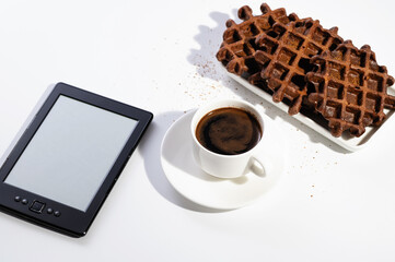 freshly brewed black natural coffee in a cup and with chocolate waffles for breakfast on a white background in a sunny morning, reading on an electronic reader