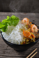 A plate of rice noodles stands on an old wooden table top. The side dish consists of fried onions, carrots, paprika, shrimps. Vertical