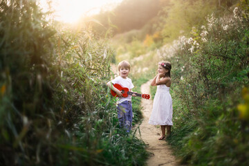 Fototapeta premium Cute romantic children a couple, a boy plays the guitar and sings, a girl in a dress is dancing. Children in a summer park barefoot on the grass. happy carefree childhood and romance