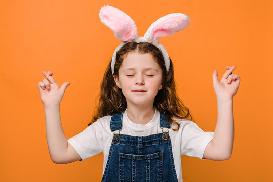 Close up portrait of carefree charming preschool girl kid cross fingers and closing eyes, wears pink bunny fluffy ears, isolated on orange background studio. Easter holiday and making wish concept