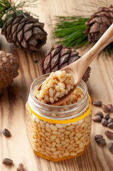 Pine nut with honey in a transparent jar. The jar is open. On the jar is a wooden spoon with honey and nuts. Nearby are pine nuts, cones and needles. Place for a label. Close-up. Light background.