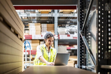 Asian female worker in safety vest sitting and working with computer laptop in storage warehouse. people, warehouse and industry concept