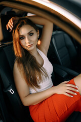 Fototapeta na wymiar A beautiful young girl sits in a car in the evening in the sunset summer sun in an empty parking lot
