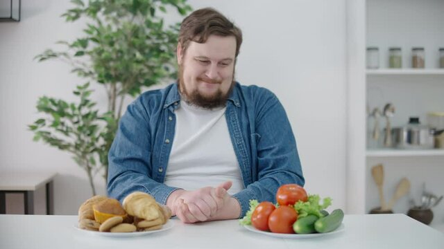 Hesitating plump man looking at muffins and vegetables on table, food choice