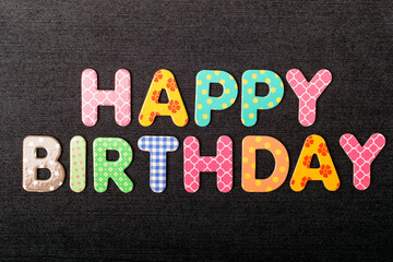 Card with Happy Birthday words made from mixed vivid colored wooden letters on a textured dark black textile material that can be used as a message.