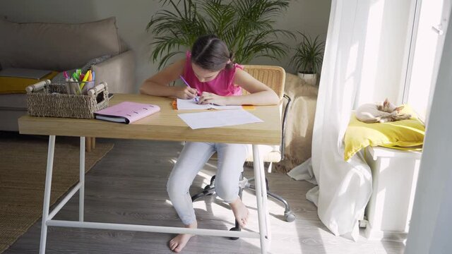 Teenager girl in pink blouse draws in paper notebook with marker leaned over the bamboo table near window in light spacious room on sunny day. schoolgirl studies at home and does school homework