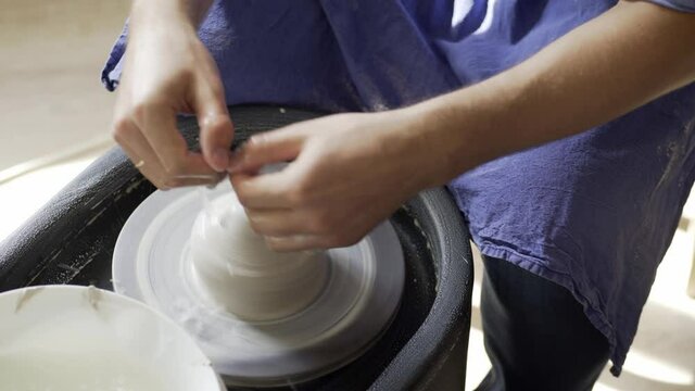 Upper view unrecognizable man holds hands on rotating clay piece forming pottery item on potters wheel and washes in bowl at workshop in ceramic studio close-up