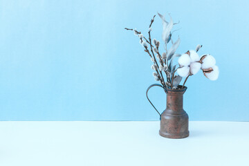 Still life with a small copper jug with a bouquet of dried flowers on a white table with a blue wall. Cotton flowers, eucalyptus, willow.