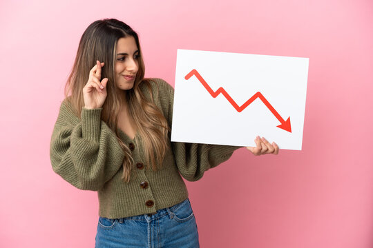 Young caucasian woman isolated on pink background holding a sign with a decreasing statistics arrow symbol with fingers crossing