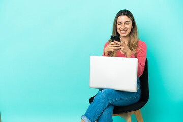 Young caucasian woman sitting on a chair with her pc isolated on blue background sending a message with the mobile