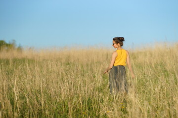 Naklejka na ściany i meble Beautiful woman in yellow shirt and long skirrt standing elegant in beige wheat field. Back view, bright blue sky