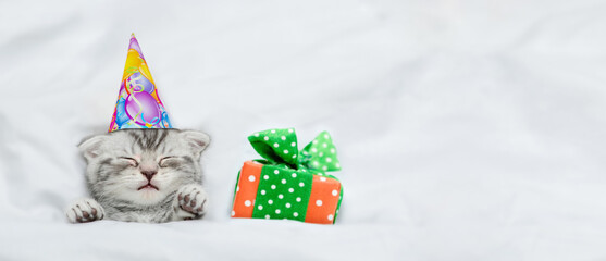Cute kitten wearing party cap sleeps with gift box under white blanket on a bed at home. Top down...