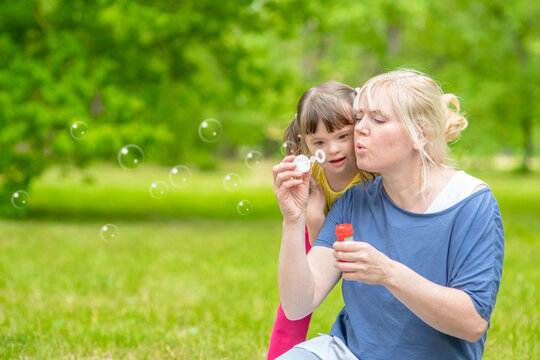 Woman and little girl with syndrome down blow bubbles in a summer park. Empty space for text