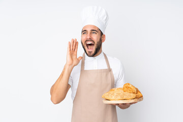 Male baker holding a table with several breads isolated on white background shouting with mouth wide open