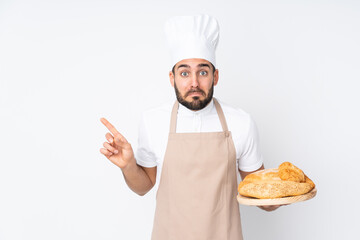 Male baker holding a table with several breads isolated on white background pointing to the laterals having doubts