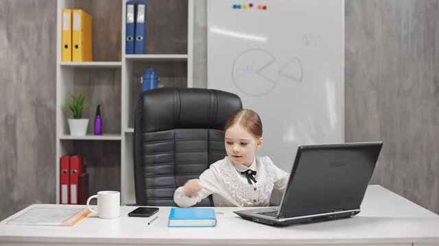 A smart, smiling little girl is talking on the phone, taking notes in the office on a laptop on a blackboard with a picture of a cake in the background. Communication in the business concept.