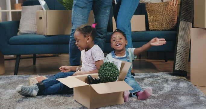 Beautiful happy smiling small two african american girls sitting on the floor with carton box full of different things near their parents in relocation day into new apartment