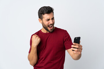 Young handsome man with beard using mobile phone isolated on white background celebrating a victory
