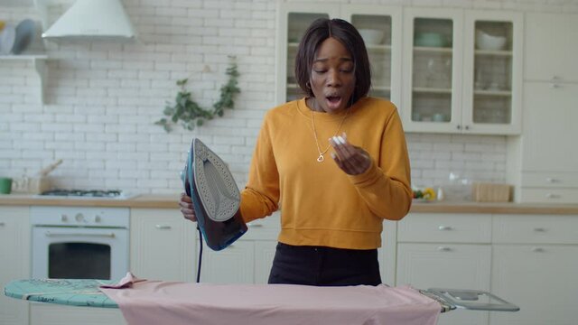 Portrait of adorable teenage black girl doing household chores, ironing clothes with iron on ironing board and getting iron burn on finger while making housework in home interior during weekend.