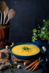 Angled image of a blue bowl of yellow carrot and coriander soup with ingredients arranged around, and rough bread, cloth and spoon, and a background vase with wooden spoons, against a blue background.