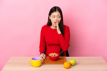 Young Chinese girl  having breakfast in a table whispering something with surprise gesture while looking to the side