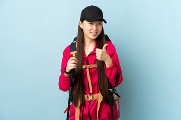 Fototapeta premium Young Chinese girl with backpack and trekking poles over isolated blue background giving a thumbs up gesture