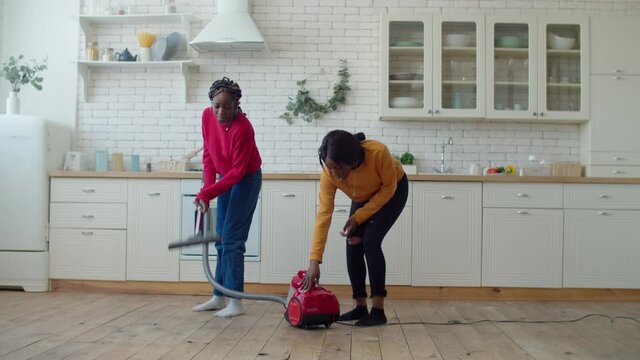 Disappointed confused lovely teenage african american sisters having problems with vacuum cleaner, trying to fix household device, expressing irritation and frustration while doing housework.