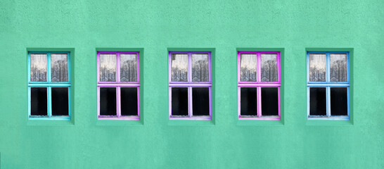 Fensterreihe mit fünf alten Fenstern, Holzrahmen bei allen mit unterschiedlichen pastellfarben angestrichen, die Hausfassade ist grün.