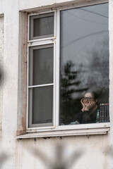 Fototapeta na wymiar A girl in a dirty window of an office building with shabby walls, leans her face in her hands, looks out the window at the street and laughs. Cloudy weather. View through the window. Street photo