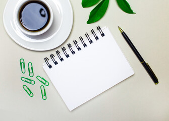 On the desktop is a white cup with coffee, a green plant and paper clips, a pen and a blank notebook with a place to insert text. Template. Flat lay. Workplace close up