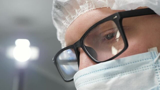 Doctor at work. Close-up of doctor male face in surgical mask and glasses, who is carefully looking down and to side, perhaps action takes place in operating room and doctor is surgeon. Slow motion