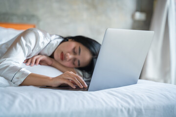 Beautiful asian girl in white pajamas sleeps at bed with laptop, laptop after work.
