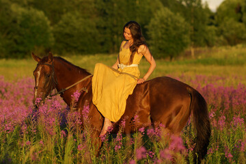 Beautiful woman with long hair in yellow dress riding bareback a brown horse in among purple flowers in green field
