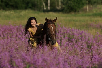 Beautiful woman with long hair in yellow dress riding bareback a brown horse in among purple flowers in green field