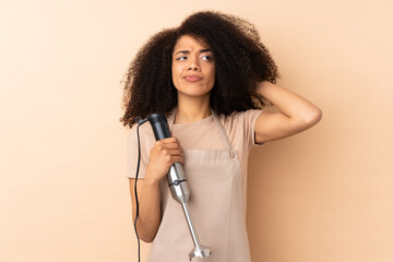 Young african american woman using hand blender isolated on beige background having doubts and with confuse face expression