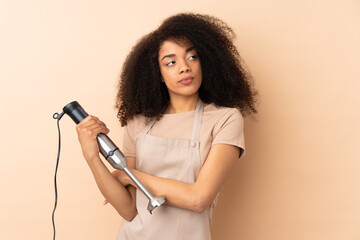 Young african american woman using hand blender isolated on beige background thinking an idea